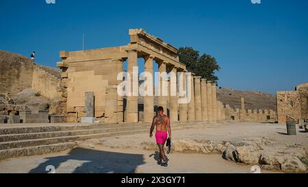 Person, die vor den beeindruckenden Ruinen eines antiken Tempels unter einem klaren blauen Himmel läuft, Akropolis von Lindos, Morgenlicht, Propyläen mit Stockfoto