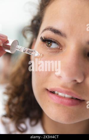 Zu Hause eine weiße Frau mit lockigem Haar, die Serum aufträgt und ein schlichtes weißes Top trägt Stockfoto