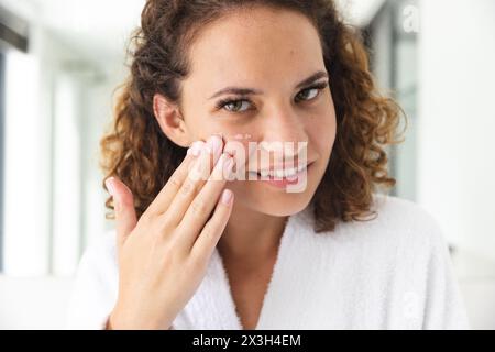 Eine junge kaukasische Frau, die zu Hause Creme auf ihr Gesicht aufträgt, im Badezimmer stehend Stockfoto