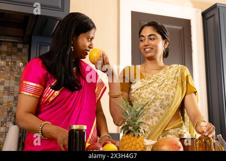Indische Mutter in rosa Sari und Teenager-Tochter in gelbem Sari, die zu Hause Essen zubereitet. Apfel halten, Mutter plaudert mit ihrer Tochter im Teenageralter, beides Stockfoto