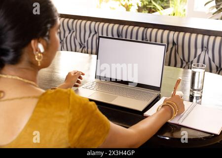 Indische junge Frau, die zu Hause auf einem Laptop schreibt, Notizbuch und Stift neben ihr, Kopierraum Stockfoto