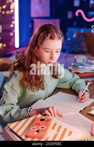 Vertikale Aufnahme eines hart arbeitenden Teenager-Mädchens, das am Schreibtisch in ihrem Zimmer sitzt und in Notizbuch schreibt, während er abends Hausaufgaben macht Stockfoto