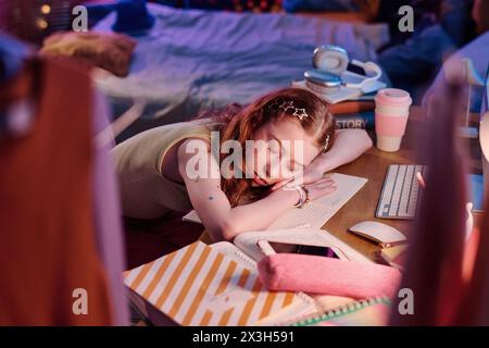 Blick aus einem hohen Winkel auf das müde rothaarige Mädchen, das beim Sitzen am Schreibtisch und bei Hausaufgaben am Abend einschlief Stockfoto