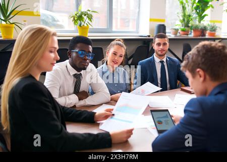 Weibliche Führungskraft leitet Geschäftstreffen mit verschiedenen Teams in modernen Büros. Junge multirassische Gruppe diskutiert Strategie, überprüft Diagramme. Professionelle Zusammenarbeit Stockfoto