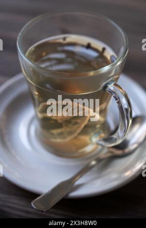 Glas gefüllt mit heißem Tee mit einer Zitronenscheibe auf einer weißen Untertasse mit einem Löffel daneben auf einem Holztisch. Geringe Schärfentiefe Stockfoto