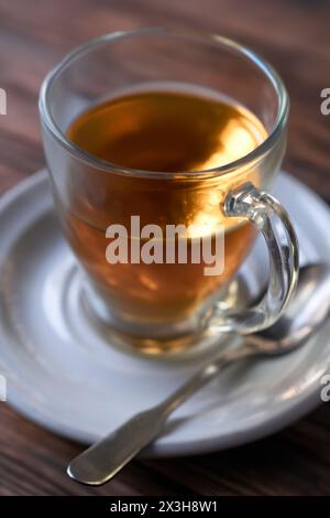 Glas gefüllt mit heißem Tee, auf einer weißen Untertasse mit einem Löffel daneben auf einem Holztisch. Geringe Schärfentiefe, Fokus auf den Griff Stockfoto