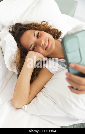 Eine junge kaukasische Frau, die ihr Smartphone hält, während sie zu Hause im Bett liegt Stockfoto