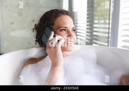 Eine junge kaukasische Frau, die sich zu Hause im Seifenbad entspannt und telefoniert Stockfoto