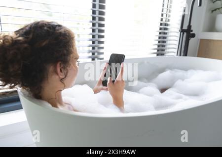 Eine junge kaukasische Frau, die sich zu Hause im Schaumbad entspannt, ein Smartphone hält, einen Kopierraum Stockfoto