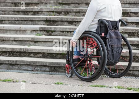 Frau im Rollstuhl hält an und wartet unten auf einer unzugänglichen Straßentreppe, die nicht hinaufgehen kann. Schwierigkeiten- und Behindertenkonzepte. Stockfoto