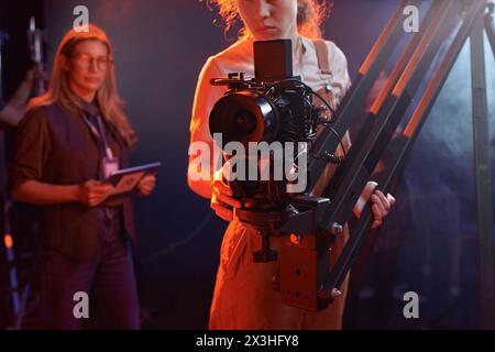 Zugeschnittene Aufnahme einer jungen Frau, die eine Videokamera auf einem Bohrfahrzeug im Studio mit Neonlichtern im Kopierraum bedient Stockfoto