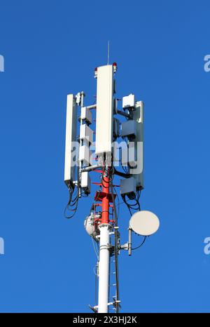Mobiltelefonsender und -Antennen in verschiedenen Formen und Größen, die auf einem hohen starken roten und weißen Metallmast auf klarem blauem Hintergrund montiert sind Stockfoto