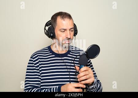 Bärtiger hispanischer Mann in seinen 40ern, der einen gestreiften Pullover trug und verwirrt aussah, während er ein Mikrofon und Kopfhörer inspizierte, isoliert auf beigefarbenem Hintergrund. Stockfoto