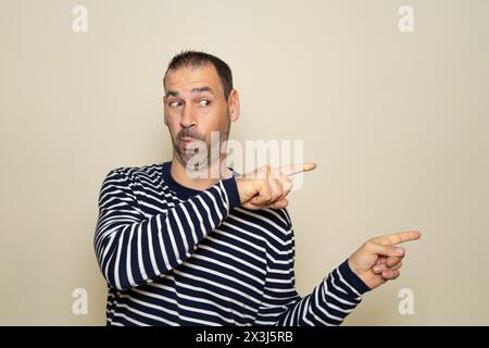 Bärtiger hispanischer Mann in seinen 40ern, der einen gestreiften Pullover trug, der mit den Zeigefingern zur Seite zeigt, mit einem Ausdruck der Überraschung. Isoliert am Stockfoto