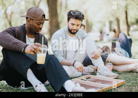 Multirassische Freunde spielen an einem sonnigen Tag Backgammon in einem Park und genießen ein unbeschwertes Treffen im Freien Stockfoto