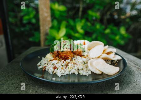 Nasi Goreng mit Schweinefleisch, Gemüse, Chili und Crackern auf einem schwarzen Teller auf dem Steintisch Stockfoto