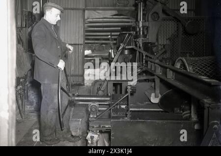 1940er Jahre, historischer, männlicher Arbeiter, turing großes Metallrad, um Maschinen aufzuwickeln, flache Kappe auf dem Kopf, Zigarette hinter dem Ohr. Stockfoto