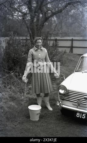 1960er Jahre, historisch, draußen auf einem Hinterrasen, eine Frau, die ein kleines Auto aus der Zeit putzt, eine Austin A40. Der A40 wurde 1958 von der Britsh Motor Corporation (BMC) hergestellt und von der italienischen Firma Pininfarina gestaltet. Der Wagen war bis 1967 mit verschiedenen Modellen in Produktion. Stockfoto