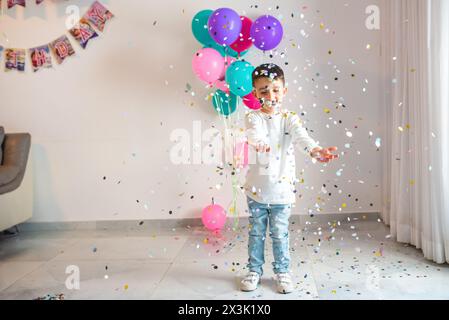 Freude erfüllt die Luft, während ein kleines Kind auf einer Geburtstagsfeier mit bunten Konfetti und Ballons spielt. Der Junge strahlt vor Freude in einem bunten Konfetti, bei seiner Geburtstagsfeier. Stockfoto