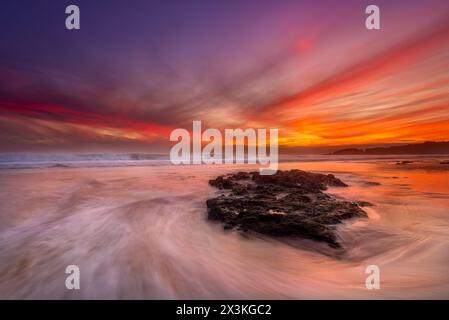 Wellen umgeben einen typischen Felsen an einem Strand in der Algarve in Portugal bei Sonnenuntergang Stockfoto