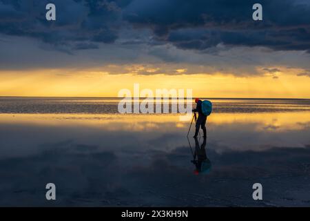 Ein Fotograf mit Stativ und Kamera steht auf einer ruhigen, reflektierenden Seeoberfläche bei einem atemberaubenden Sonnenuntergang, der Ruhe und Gelassenheit ausstrahlt. Stockfoto