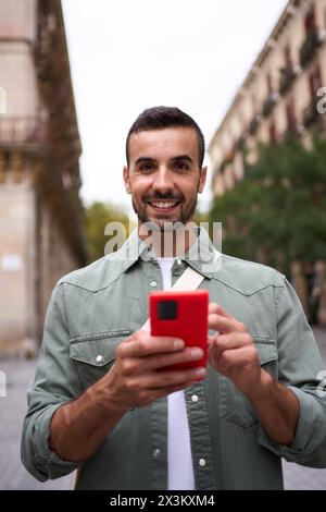 Porträt junger europäischer Mann steht und sieht lächelnd in die Kamera, die ein Handy in Madrid hält. Stockfoto