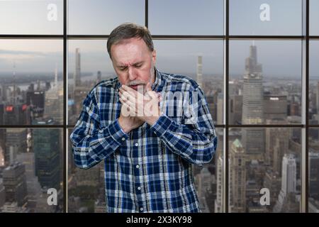 Trauriger reifer kaukasischer Mann hustet drinnen. Windows-Hintergrund mit Stadtbild-Ansicht. Stockfoto