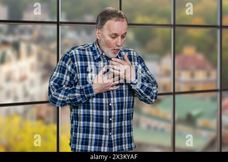 Porträt eines reifen Mannes, der drinnen hustet. Windows-Hintergrund mit Stadtbild-Ansicht. Stockfoto