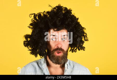 Porträt eines überraschten bärtigen Mannes in schwarzer Perücke. Partyzeit. Stilvolles Männermodell mit Bart und Schnurrbart in lockiger Perücke. Modekonzept. Schöner Hipster Stockfoto