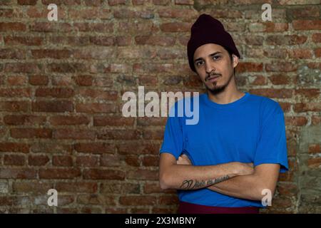 Junger Latino-Mann (28) mit überkreuzten Armen blickt direkt in die Kamera. Kopierraum, Backsteinhintergrund. Stockfoto
