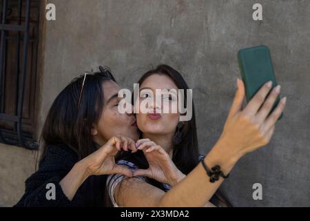 Lateinamerikanische Mutter (50) und Tochter (24) machen ein Selfie, beide machen mit ihren Händen ein Herzsymbol, während ihre Mutter ihre Tochter Zuneigung küsst Stockfoto