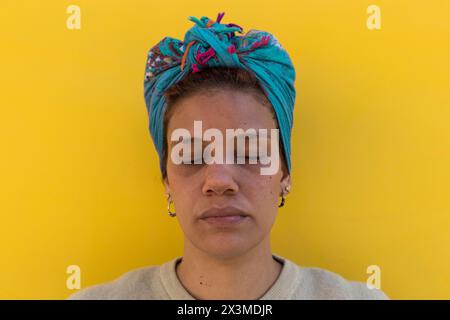 Afro-lateinamerikanische Frau (34) mit grünen Augen, Kopftuch und isoliert auf gelbem Hintergrund mit geschlossenen Augen vor der Kamera. Stockfoto