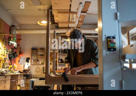 Fröhlicher Mann Zimmermann, der mit handgemachtem Tisch arbeitet. Schreiner fühlt sich mit dem Handwerksprozess zufrieden. Stockfoto