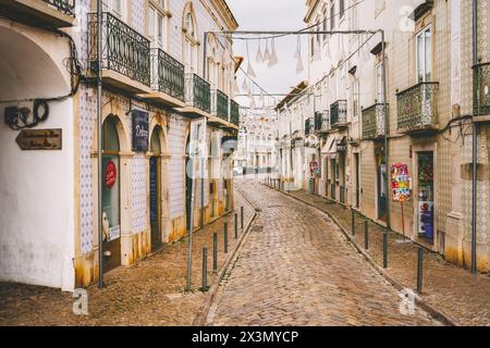 Tavira, Portugal, 8. Januar 2024. Eine kopfsteingepflasterte Straße in Tavira, Portugal, mit Autos, die an der Seite geparkt werden, und traditionellen weißen Gebäuden entlang der Straße Stockfoto