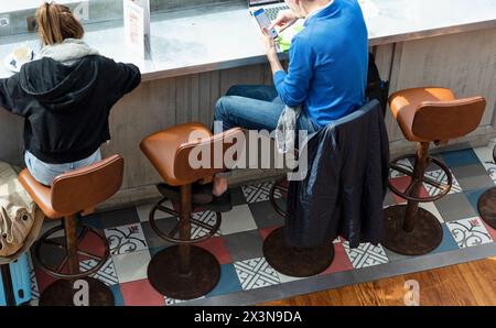 Zwei Reisende an einer Bar, die auf Lederhockern neben einer Holztheke sitzen, warten mit ihren Trolleys auf den Abflug ihres Fluges vom Flughafen CDG in Stockfoto