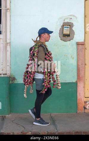 Ein Straßenverkäufer, der vor einer verwitterten Mauer läuft und Zwiebeln und Knoblauch in den Nebenstraßen der Altstadt, Trinidad, Kuba, trägt. Stockfoto