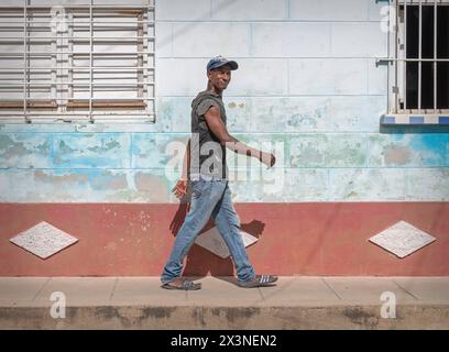 Ein glücklicher junger Mann, der auf dem Bürgersteig vor einer verwitterten Mauer spaziert, mit verblasstem Lack in den Seitenstraßen der Altstadt, Trinidad, Kuba Stockfoto