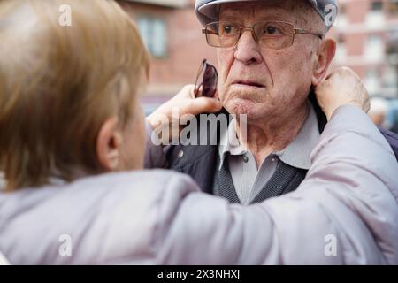 Eine alte Frau hilft seinem Mann, sich draußen anzuziehen. Abhängigkeit und Betreuung im Alter Stockfoto