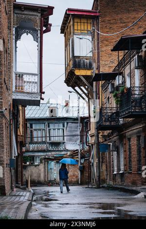 Frau mit einem blauen Regenschirm, die unter dem Regen in der Straße der Altstadt von Tiflis (Georgien) läuft, umgeben von baufälligen Häusern und Holzbalkonen Stockfoto