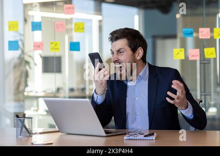 Wütender Boss-Täter, der am Telefon spricht, Mann in einem Business-Anzug schreien, Geschäftsmann am Arbeitsplatz streitet um Gespräche, arbeitet im Büro am Arbeitsplatz mit Laptop. Stockfoto