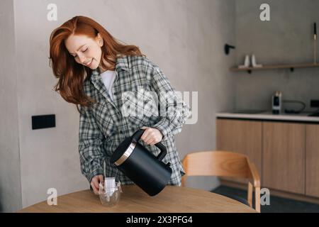 Attraktive, lächelnde junge Frau, die morgens Kaffee mit Tropfkaffeesack in einer transparenten Glasschale macht, die am Tisch steht. Vorbereitung frisch verarbeiten Stockfoto