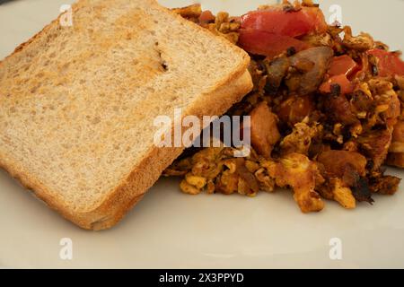 Eine Portion hausgemachter Mahlzeit mit Wurst, Zwiebeln, Tomaten und Rührei mit einer gerösteten Vollkornscheibe Brot. Stockfoto