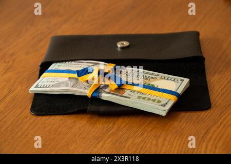 Ein Band in Form der Flagge der Ukraine, gebunden mit einer Schleife auf einem Stapel von hundert Dollarscheine liegt in einer Geldbörse auf dem Tisch, finanzielle Hilfe Stockfoto