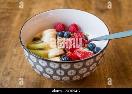 Schüssel mit frischem Joghurt (Joghurt) mit Müsli, gemischten Sommerbeeren, Banane und Kiwi mit einem Löffel auf einem Holztisch Stockfoto