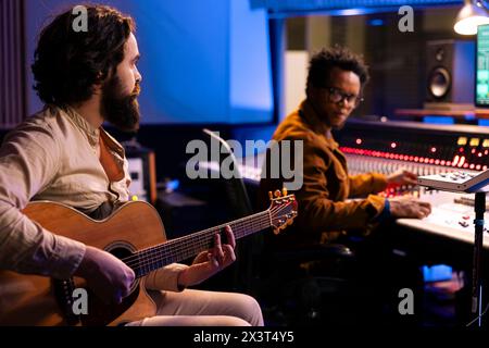 Junger Sänger Komponist kreiert einen neuen Track auf der Gitarre im Aufnahmestudio, Sounddesigner hört Musik, bevor er die Beats auf der Audiokonsole mischt. Geschickter Künstler, der Lieder im Kontrollraum komponiert. Stockfoto