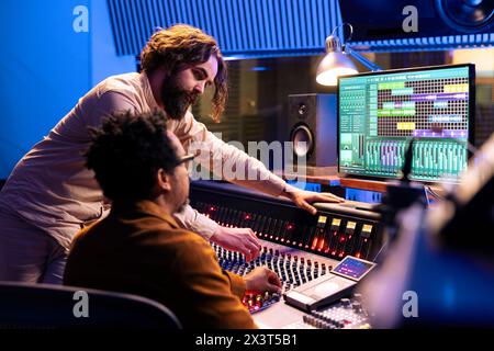 Ein erfahrener Künstler arbeitet mit einem Tontechniker zusammen, um aufgezeichnete Songs im Kontrollraum zu bearbeiten, indem er Schieberegler und Hebel auf der Platine drückt. Ein Team aus Sänger und Sounddesigner erstellt Aufnahmen im Studio. Stockfoto