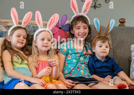 Eine Familie mit drei Mädchen, Schwestern und einem Kleinkind, Bruder sitzt auf einer Couch, zeigt ihre osterhasenohren auf dem Kopf und lächelt. Stockfoto