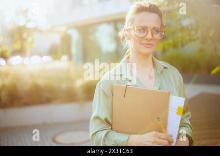 Moderne Angestellte in der Nähe des Bürogebäudes in grüner Bluse und Brille mit Dokumenten und Mappe. Stockfoto