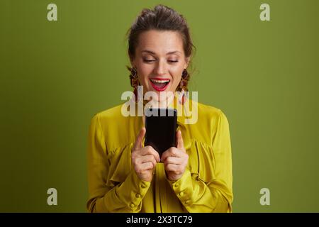 Überraschte moderne Frau isoliert auf Grün in grüner Bluse mit Smartphone. Stockfoto