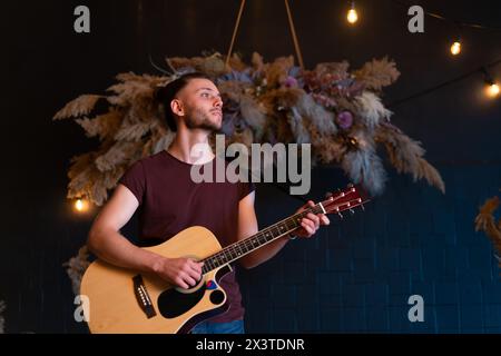 Männlicher Musiker, der akustische Gitarre spielt. Gitarrist spielt klassische Gitarre auf der Bühne im Konzert hübsch junge Hipster männlichen Gitarrist führen private Party stilvolle lange Haare Medium shoot Stockfoto